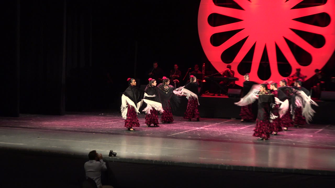 The Cabo San Lucas dance group spreads across the stage as several performers extend pale shawls in front of the red lit backdrop and seated musicians.