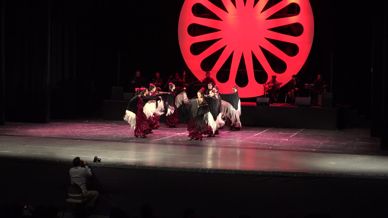 The dancers remain grouped near center stage in Cabo San Lucas, with white fringe shawls extended and the red circular backdrop glowing above the band platform.