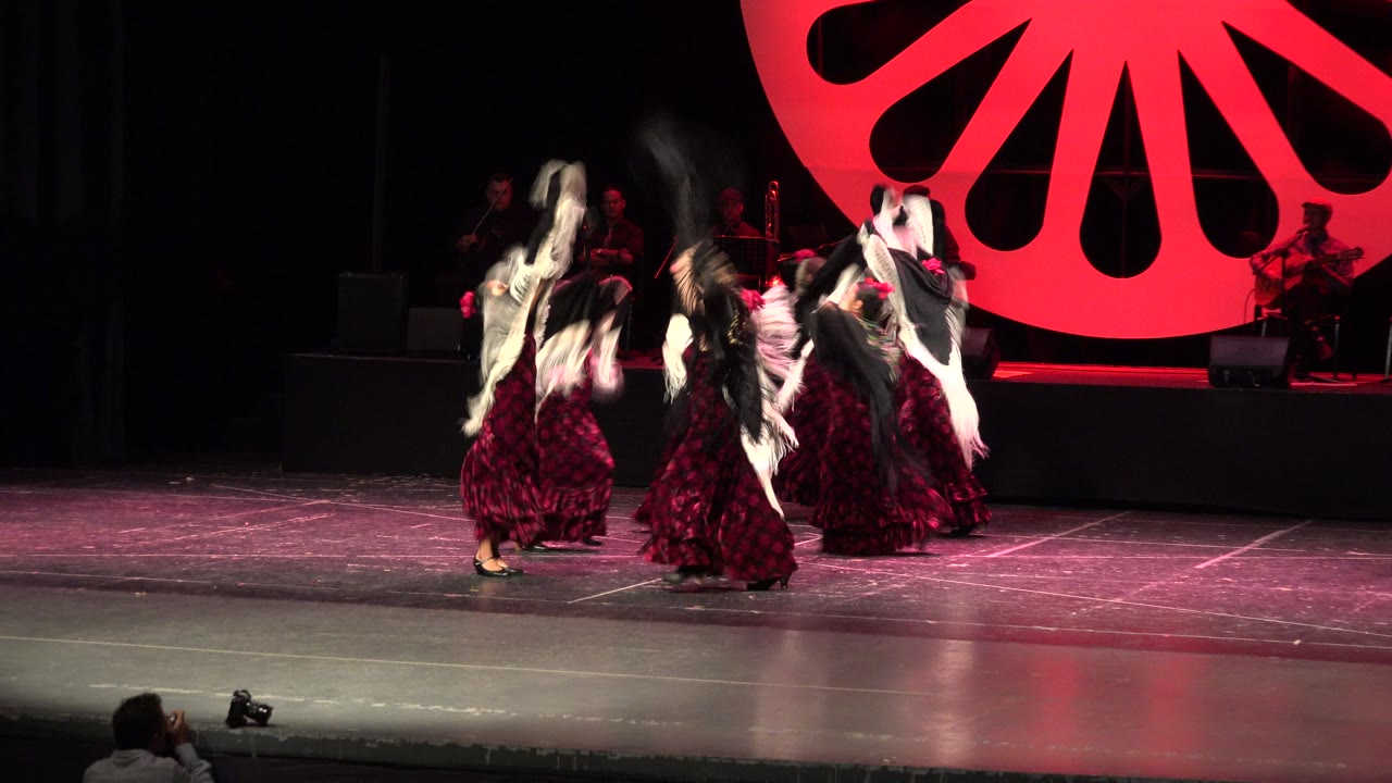 Young dancers in burgundy dresses move together on a dark stage.
