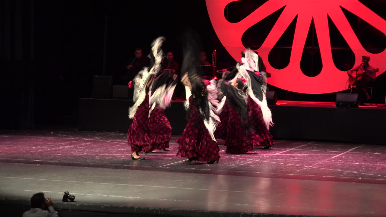 The dancers continue their Cabo San Lucas stage routine as white-fringed shawls lift above burgundy dresses, with musicians dimly visible behind them.