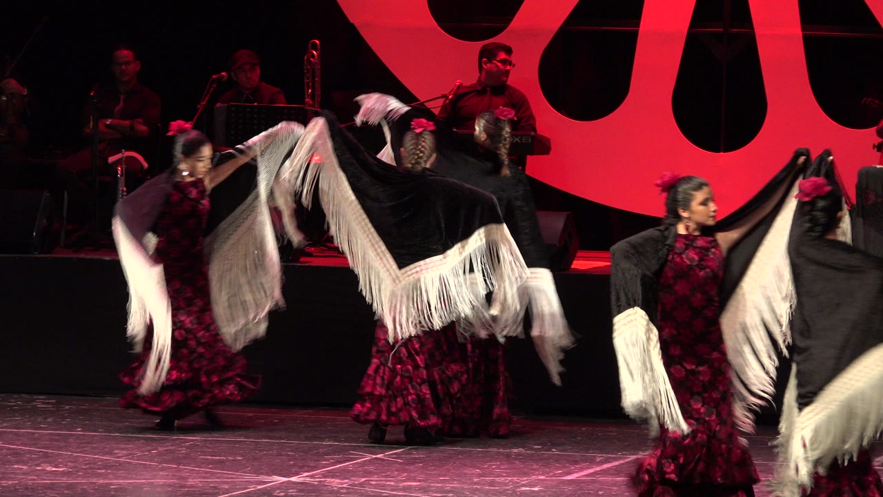 Flamenco dancers in red and black outfits wave fringed shawls on a stage.