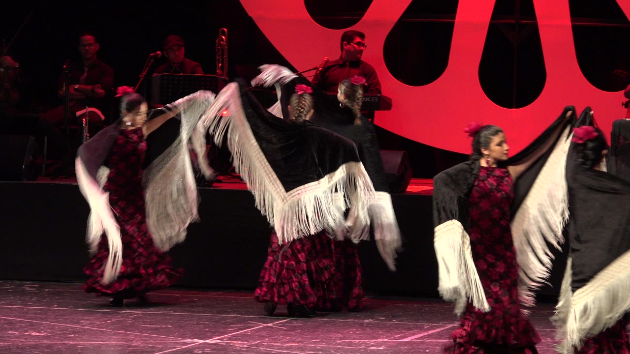 Several women in red and black flamenco dresses dance onstage in Cabo San Lucas as a central shawl creates a sweeping white fringe arc.
