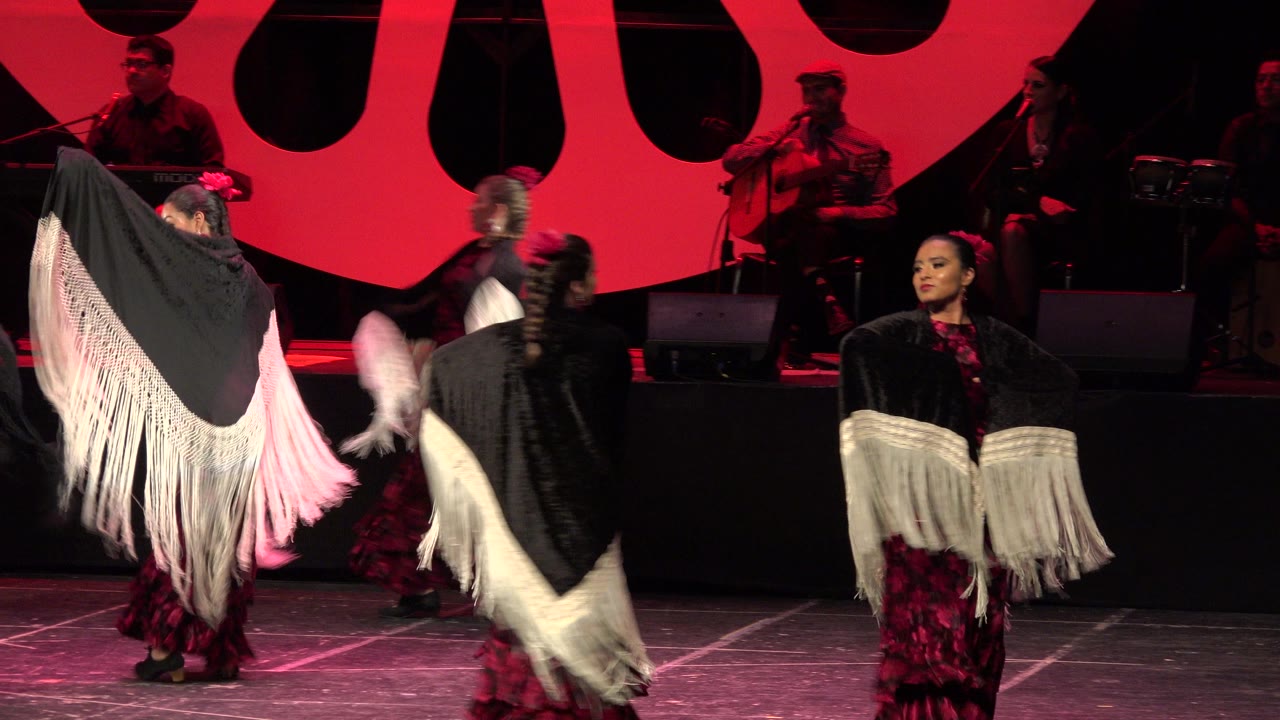 Women perform on a red-lit stage in Cabo San Lucas, Mexico, with black shawls, white fringe, red patterned dresses, and seated musicians visible behind them.