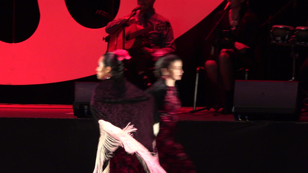 A dancer with a black shawl and white fringe passes left of another dancer in a red dress on the Cabo San Lucas performance stage, with the band partly visible behind them.
