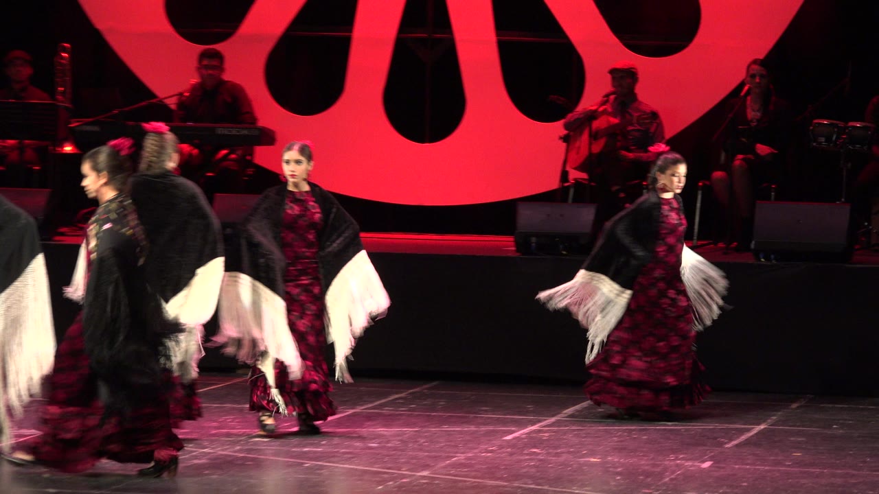 In Cabo San Lucas, Mexico, two dancers hold positions near the front as other performers pass between them under red stage light with musicians behind.