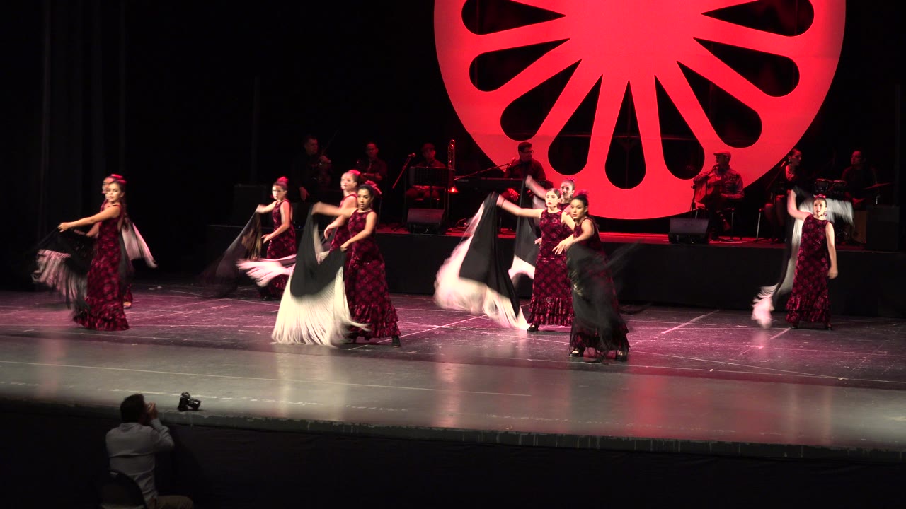 Dancers in dark red costumes continue the Cabo San Lucas stage routine, with shawls open across the center and a photographer seated near the front edge of the stage.