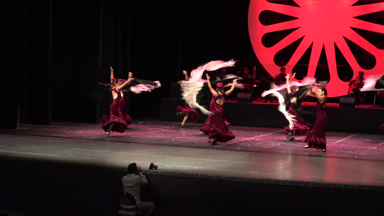 Women in red dresses dance with shawls on a theater stage.