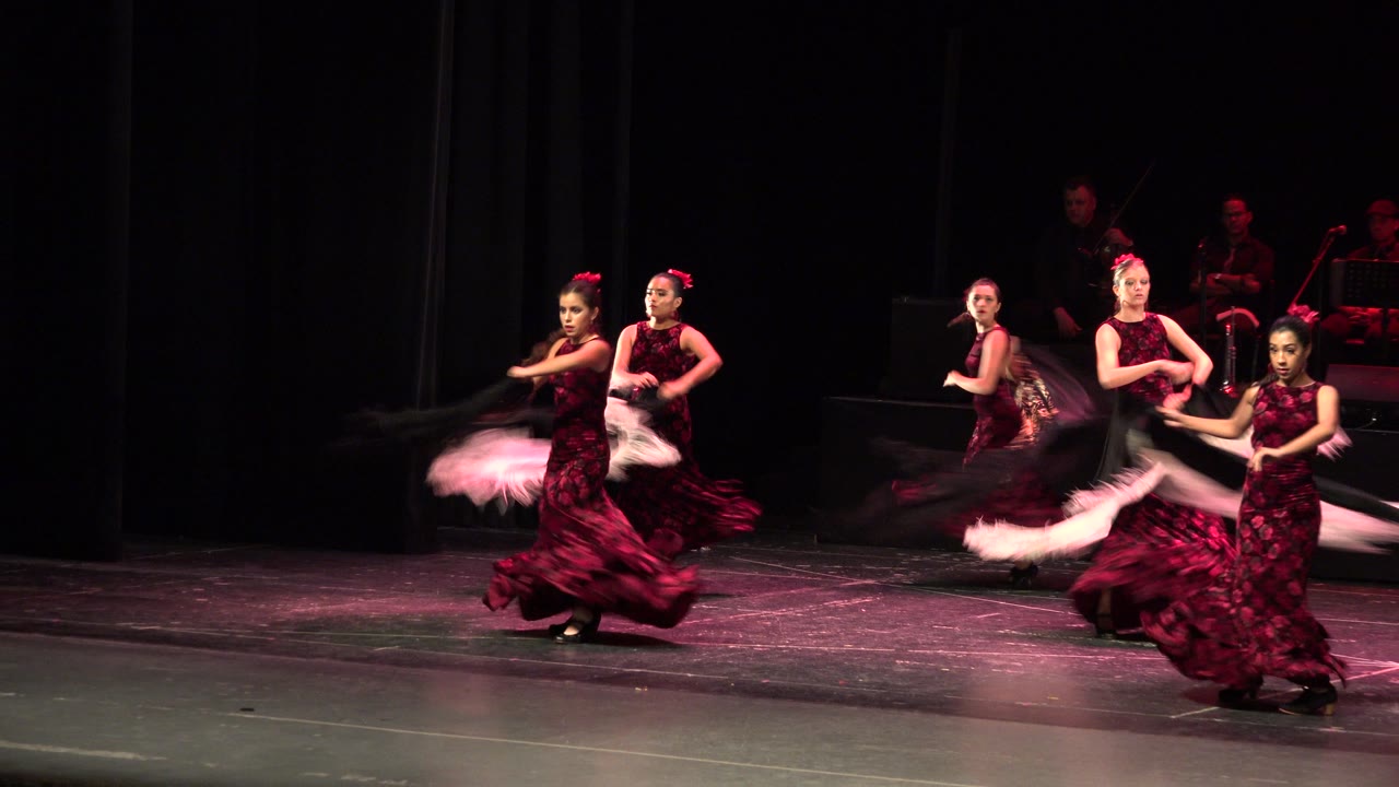 Women in red and black dresses perform on a dark stage in Cabo San Lucas, Mexico, with shawls sweeping through warm pink stage light.