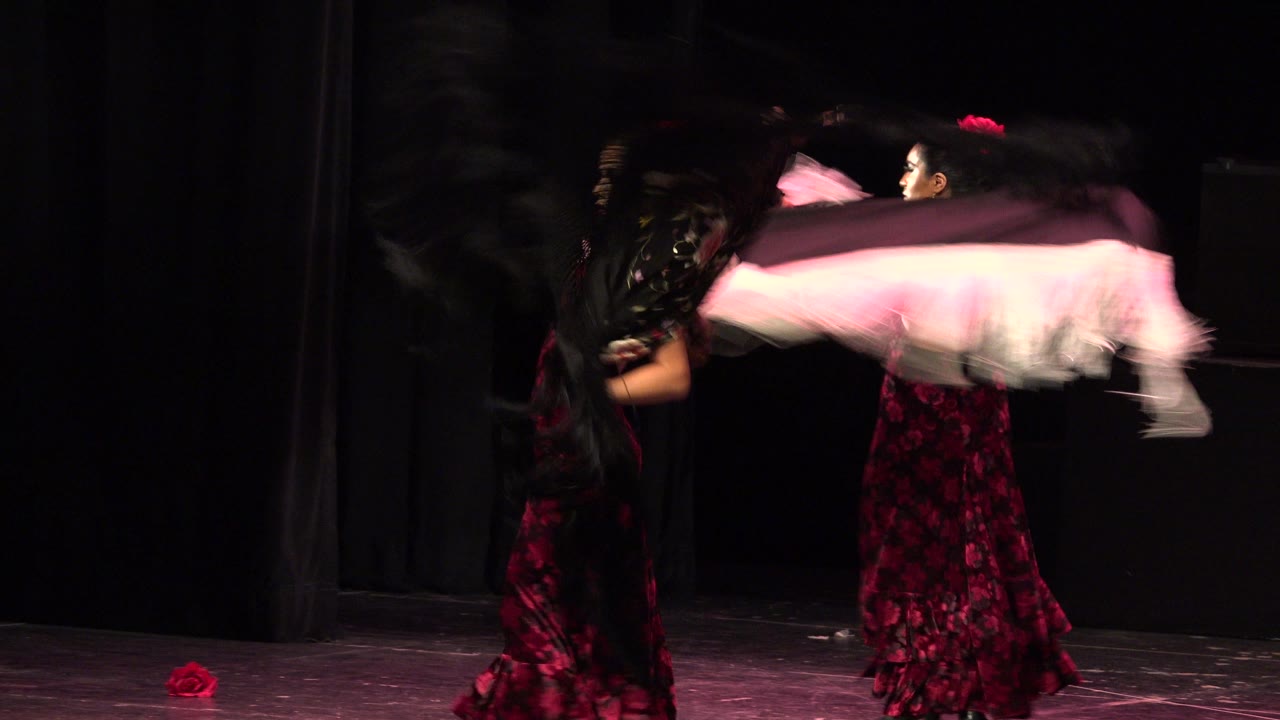 Two women in formal red and black dresses continue the Cabo San Lucas stage dance, with one pale shawl stretched horizontally and the dark backdrop behind them.