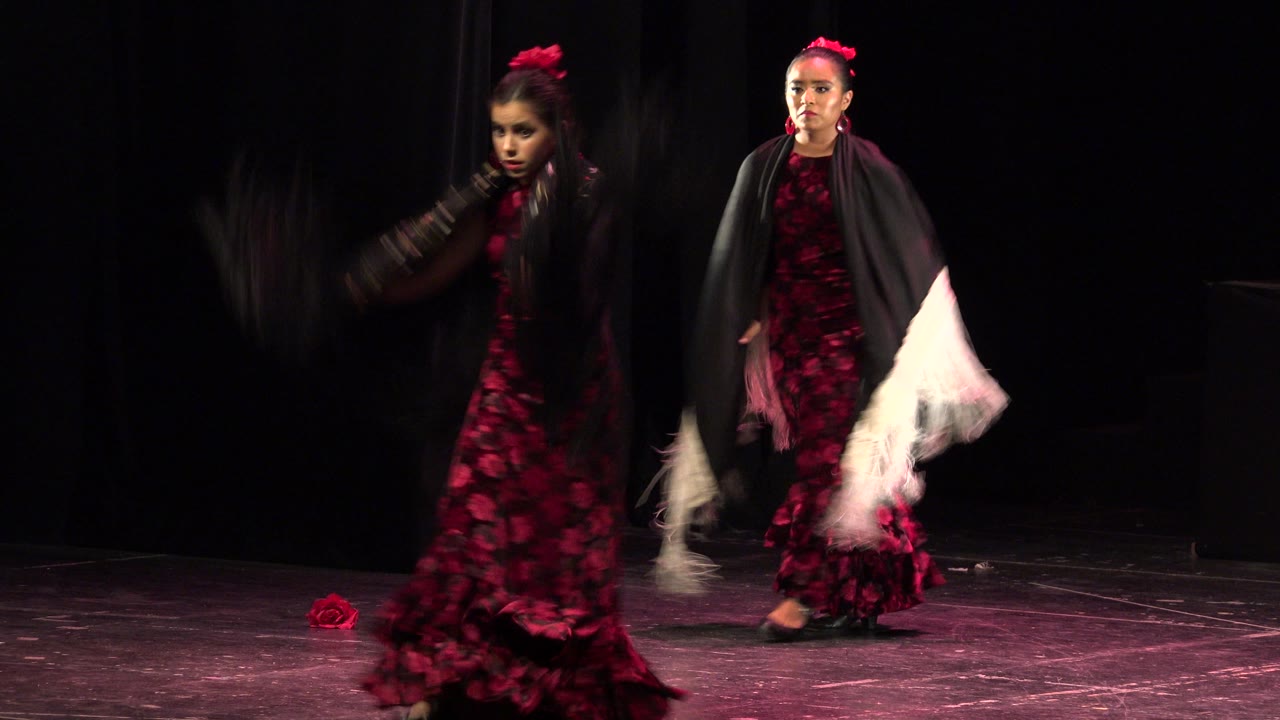 Two young dancers in red dresses continue choreography on a dark Cabo San Lucas stage, with fabric motion blur crossing the left side of the frame.