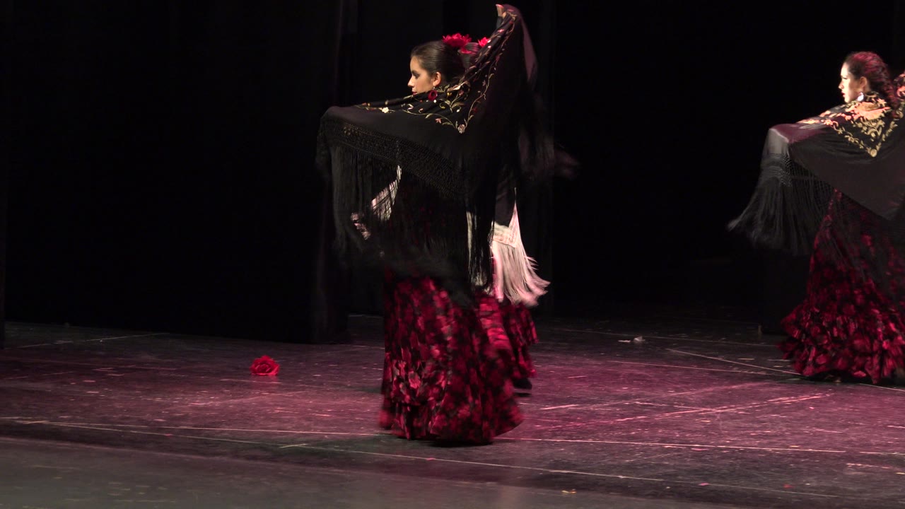 At 0:00 in Cabo San Lucas, Mexico, dancers in red-and-black patterned dresses hold black fringed shawls on a dark theater stage under pink lighting.