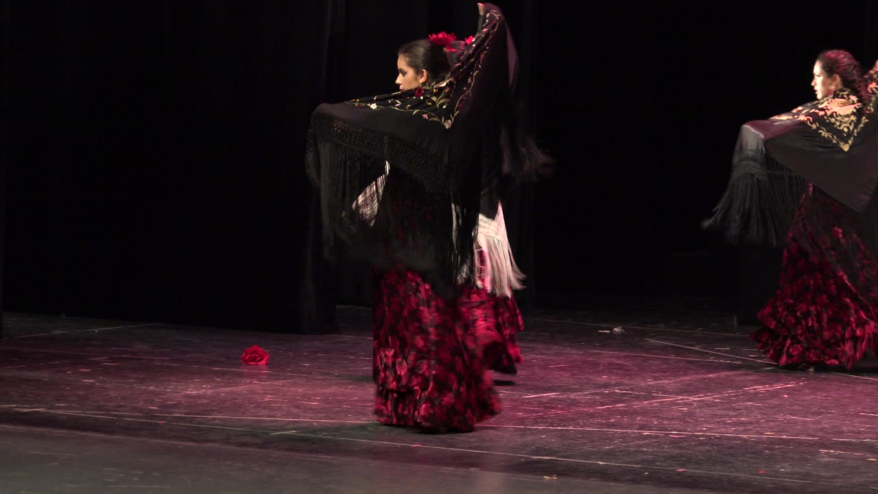 At 0:01 in Cabo San Lucas, Mexico, two dancers in red patterned dresses turn across a dark stage while black fringed shawls spread behind their arms.