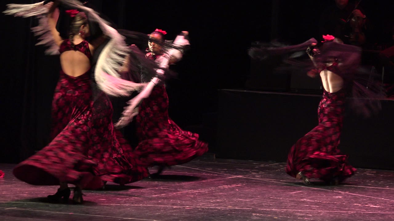 Dancers in red dresses swing shawls on a dark stage.