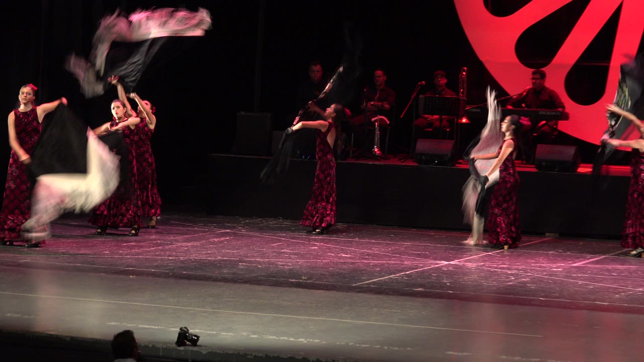 Women in purple dresses perform on a Cabo San Lucas stage as black and white fringed scarves sweep through the foreground under red lighting.