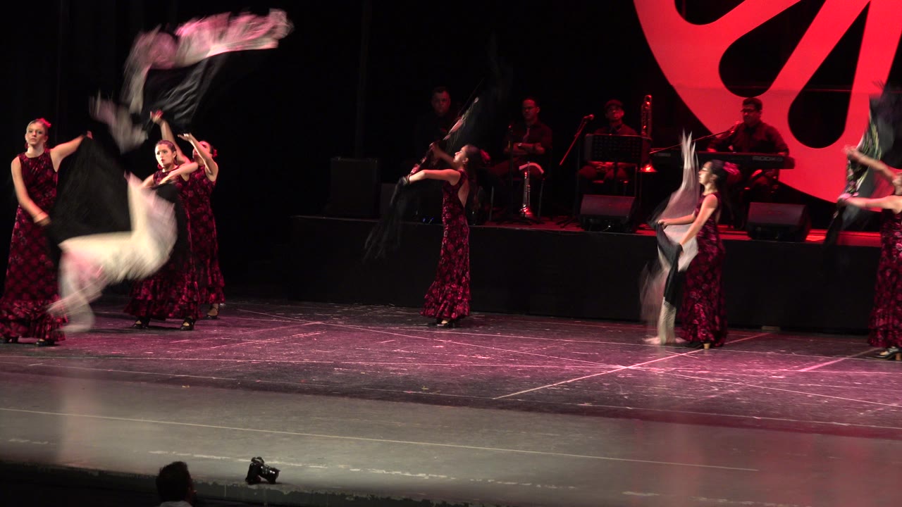 A Cabo San Lucas stage performance continues with women in purple dresses holding scarves while a large red fan-shaped scenic piece fills the background.