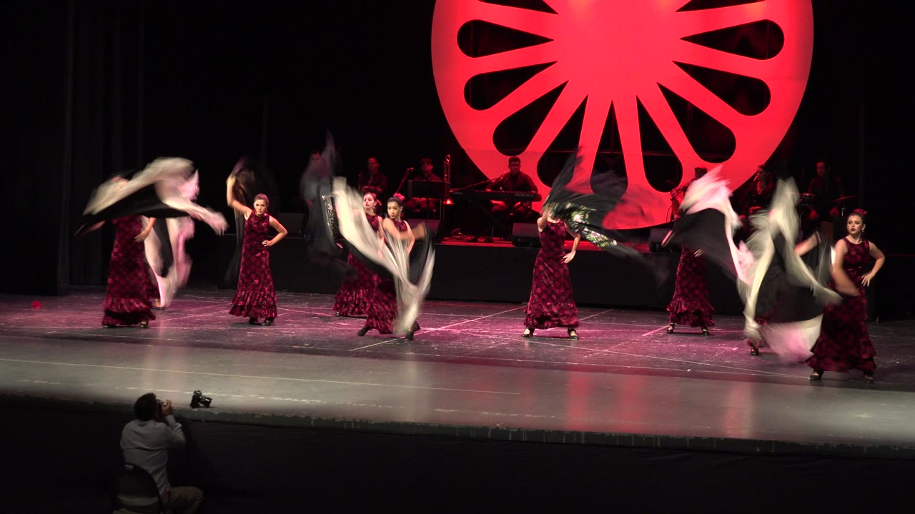 Women in red dresses dance on a black stage with shawls raised.