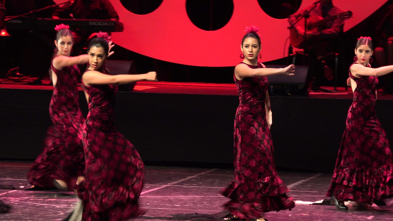 The dancers maintain a tight formation in Cabo San Lucas, with one girl facing forward while others extend their arms across the red-lit stage.