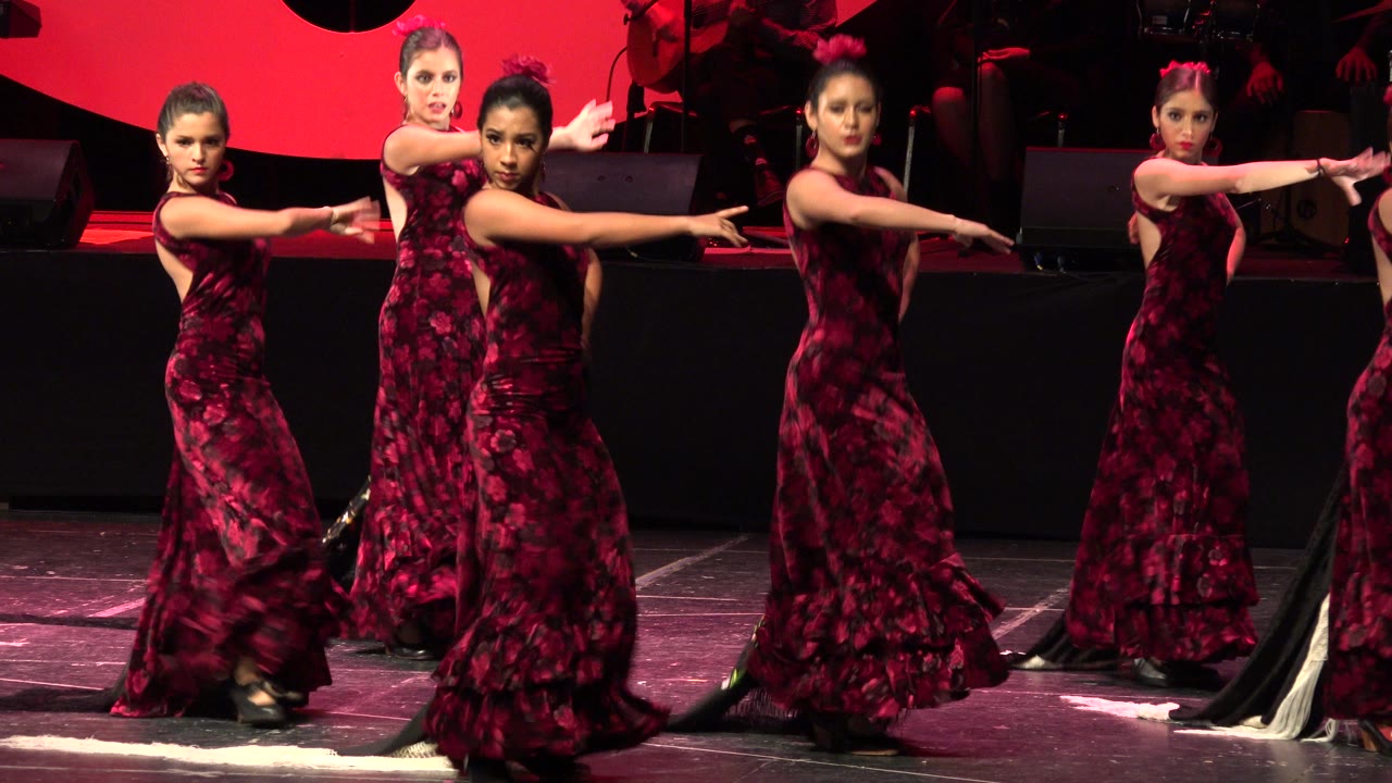 In Cabo San Lucas, Mexico, dancers in matching maroon dresses hold a forward-facing stage pose with extended arms under red lighting.