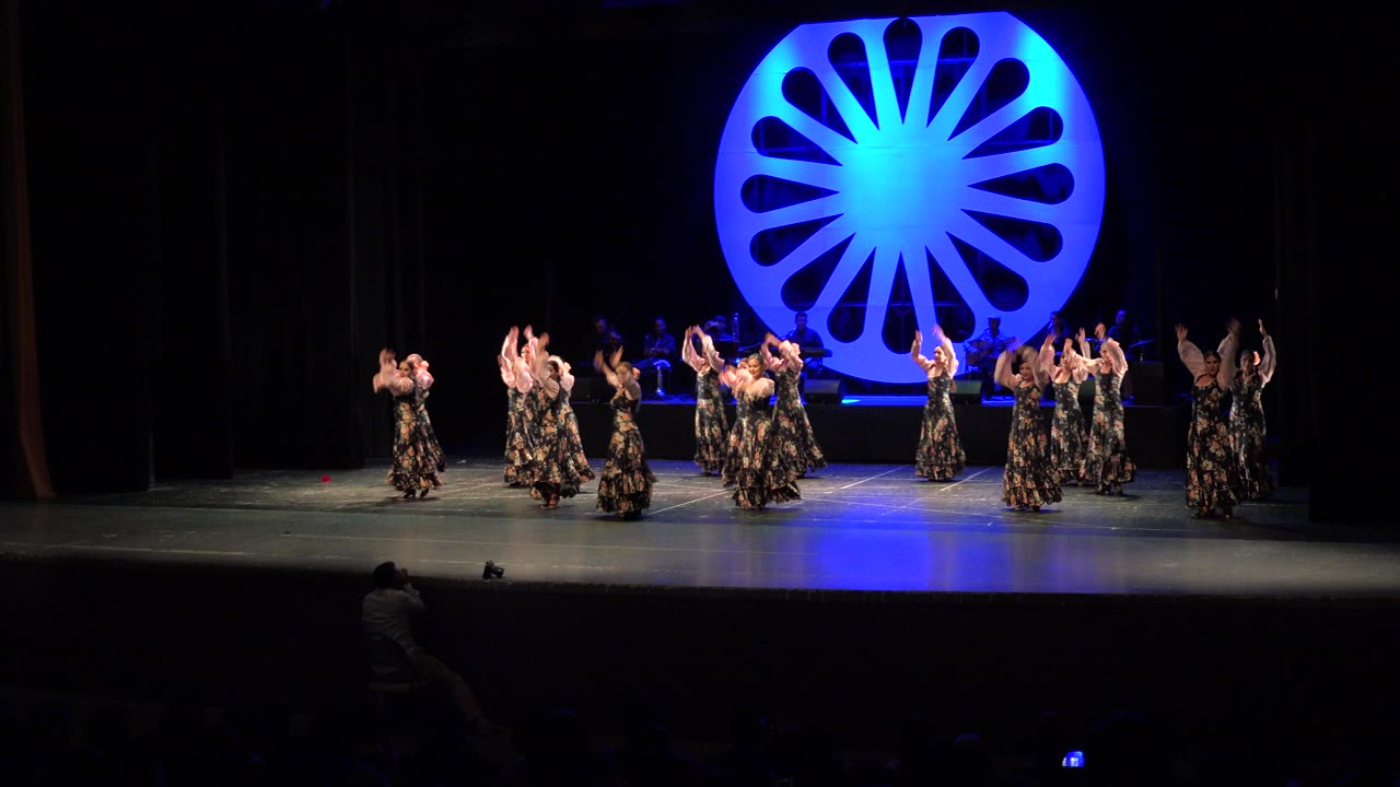 Wide frame from Cabo San Lucas, Mexico showing women in floral dresses dancing across a large stage under a bright blue circular set piece with musicians in the dark background.