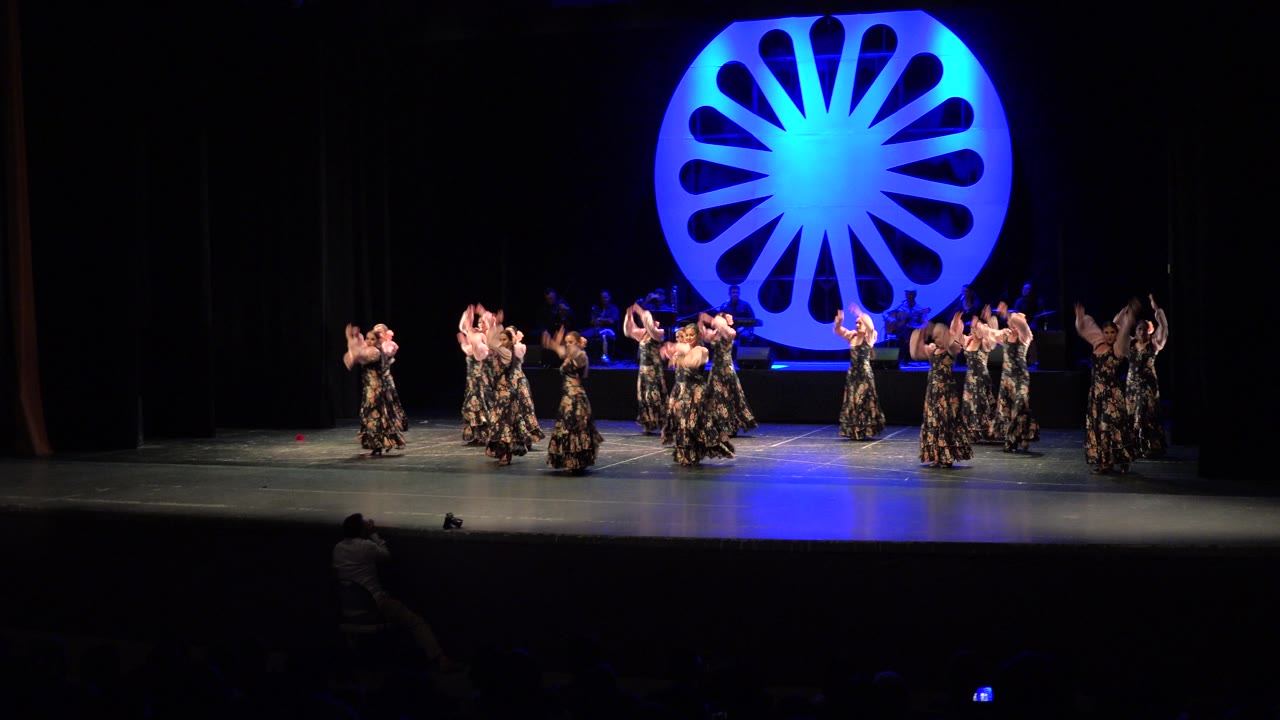 Wide stage view in Cabo San Lucas, Mexico with the ensemble clustered near center, arms raised and skirts visible under blue theatrical lighting.