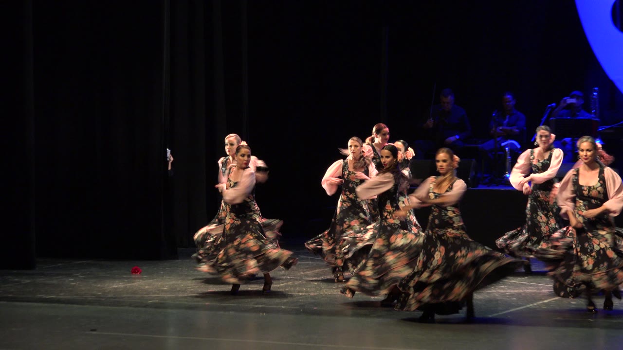 The group continues the Cabo San Lucas stage performance, with several dancers turning in floral skirts while others hold positions near the back.