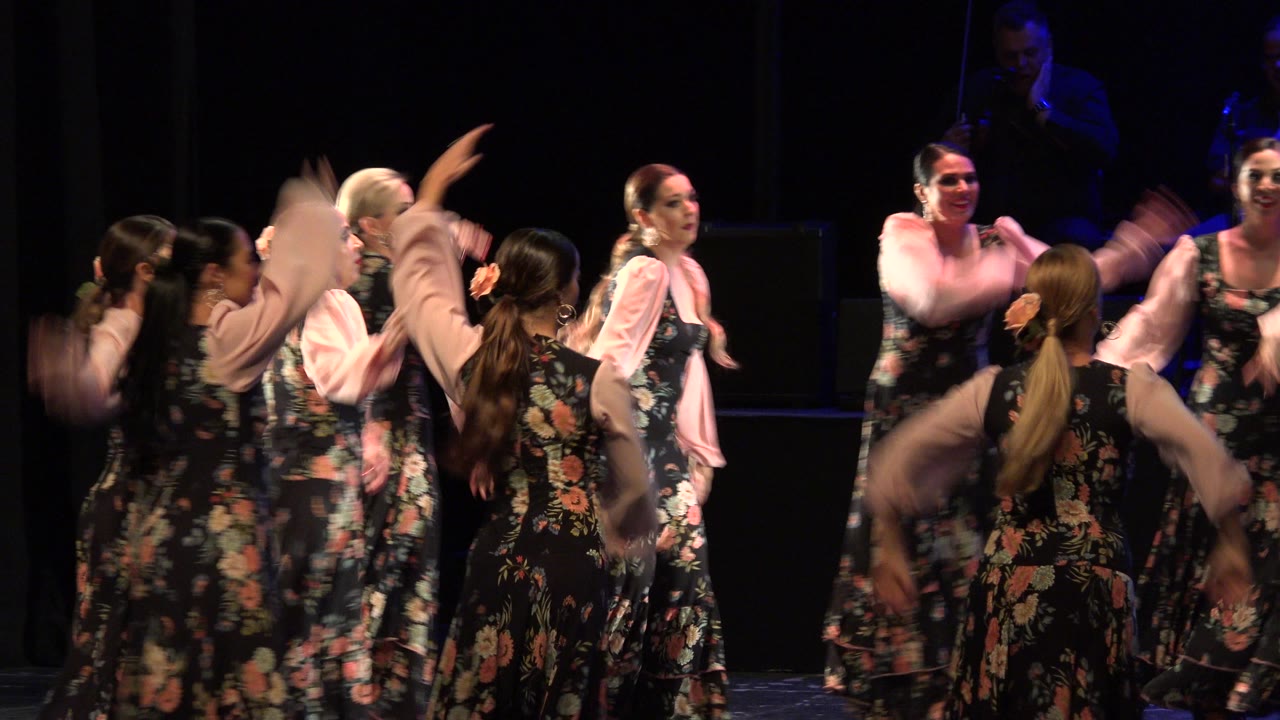 Women in black floral gowns with pale pink sleeves move in a tight group on a dark stage in Cabo San Lucas, Mexico, with faces and arms partly blurred by motion.