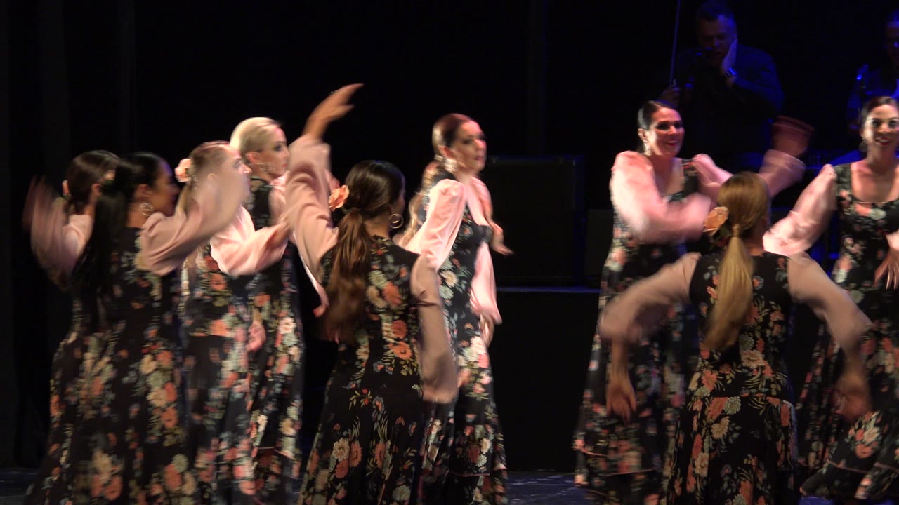 The dance group continues across the stage in Cabo San Lucas, Mexico, with raised arms, black floral skirts, pale sleeves, and blue background light behind the performers.