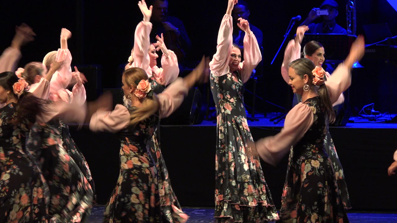 Teen dancers in floral dresses raise their arms on a dark stage.