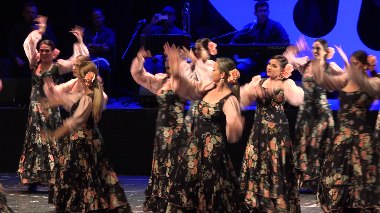 Female dancers in floral dresses perform on a blue-lit stage.