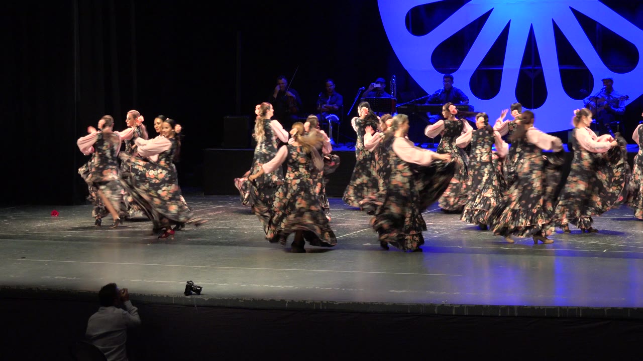 A wide stage view in Cabo San Lucas shows a group of dancers in dark floral costumes moving together while musicians sit behind a blue-lit circular backdrop.