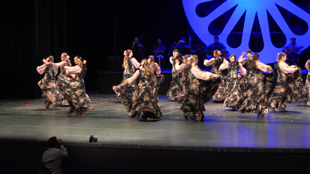 The performers remain in a wide formation on the Cabo San Lucas stage, with skirts in motion, blue light on the floor, and the band visible in the dark background.