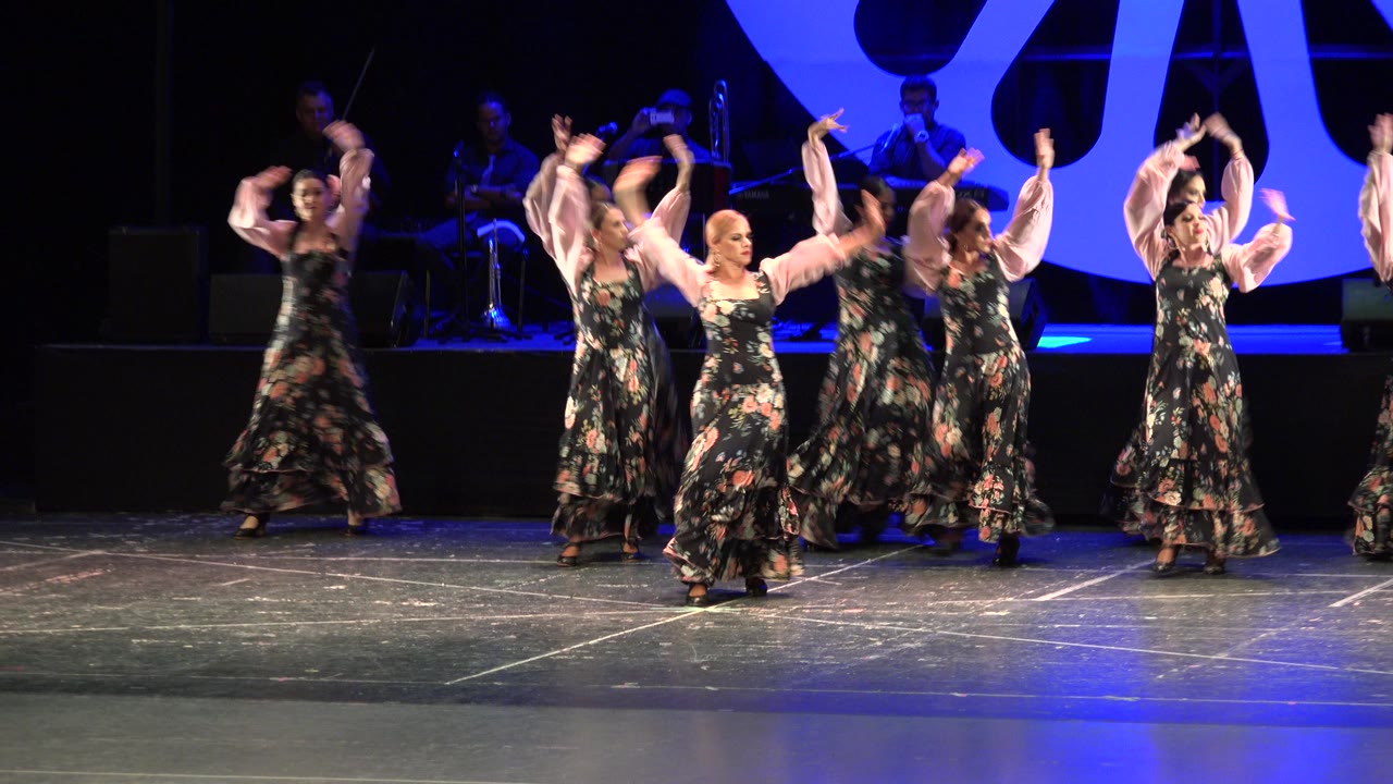 Women in floral dresses dance on a blue-lit stage.