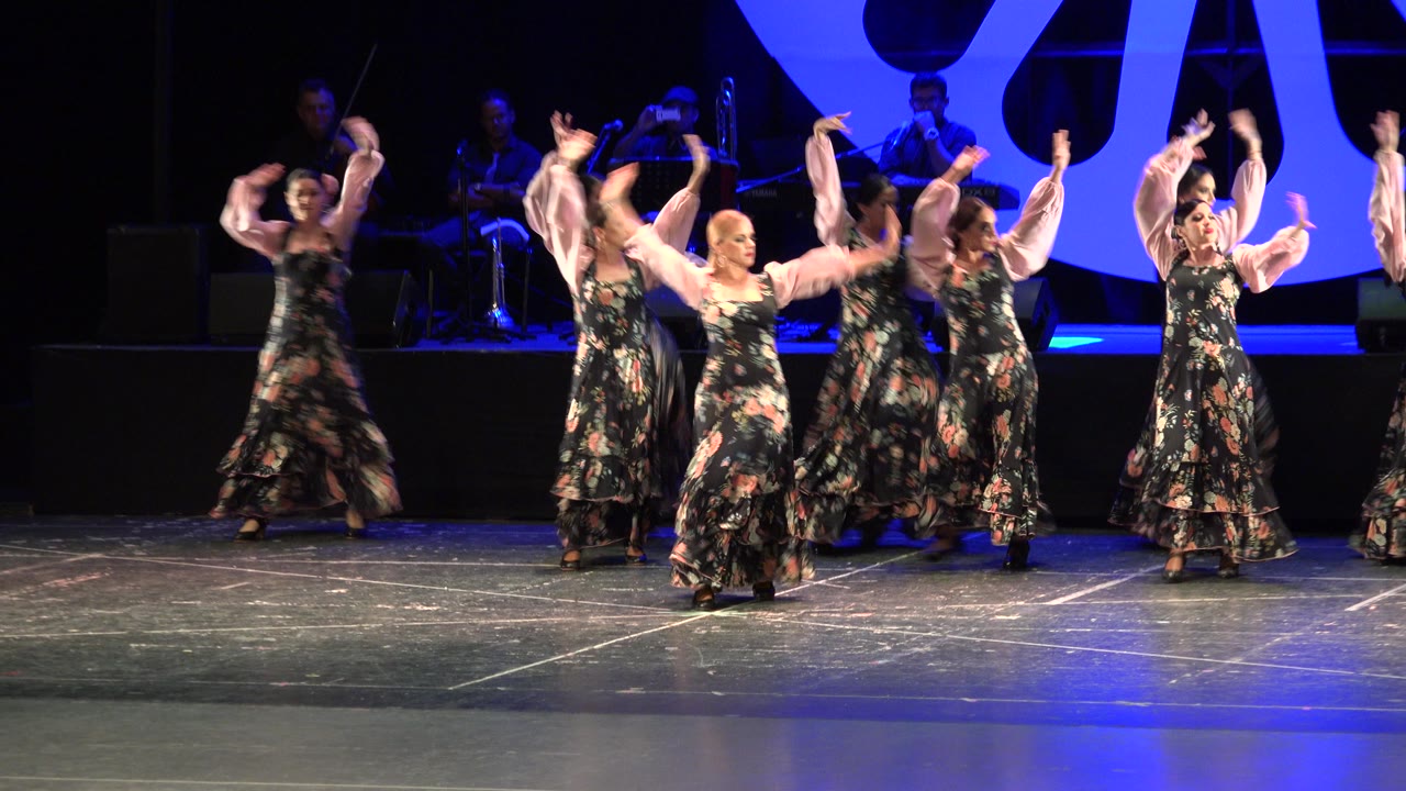 Adult women continue the choreographed dance in Cabo San Lucas, Mexico, with several raised arms, floral skirts, and a dark band area in the background.