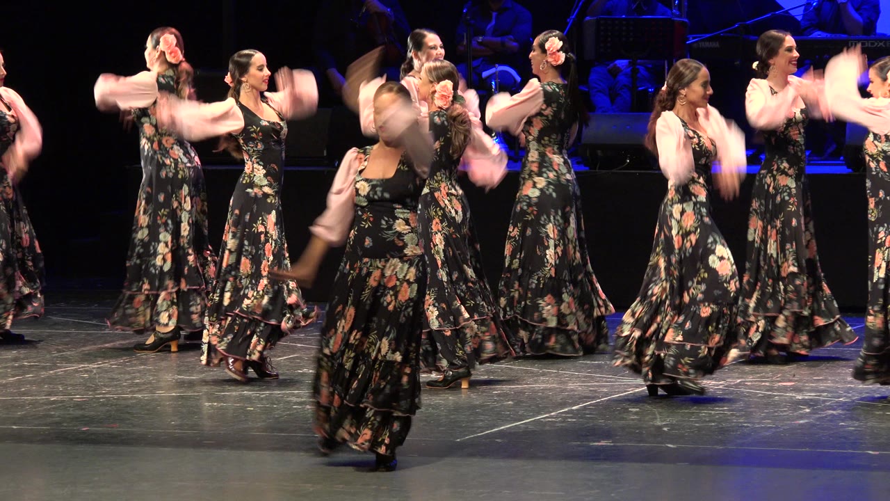 Women in floral dresses dance on a dark stage.