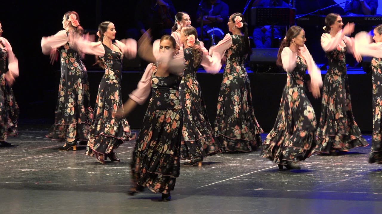 A dancer moves across the foreground with motion blur while other adult women in matching floral dresses continue the performance on a dark stage in Cabo San Lucas, Mexico.