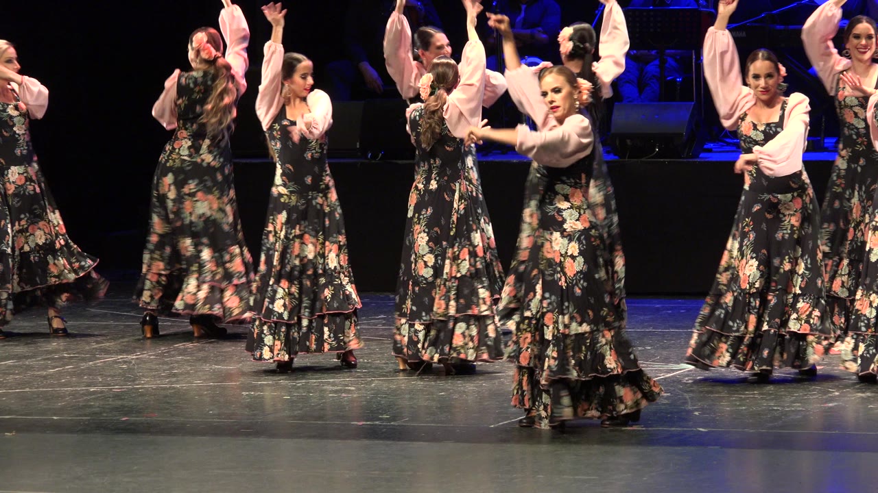 Women in floral flamenco dresses dancing on a theater stage.