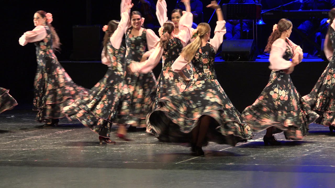A line of adult women dancers moves across the stage in matching floral dresses during the Cabo San Lucas performance.