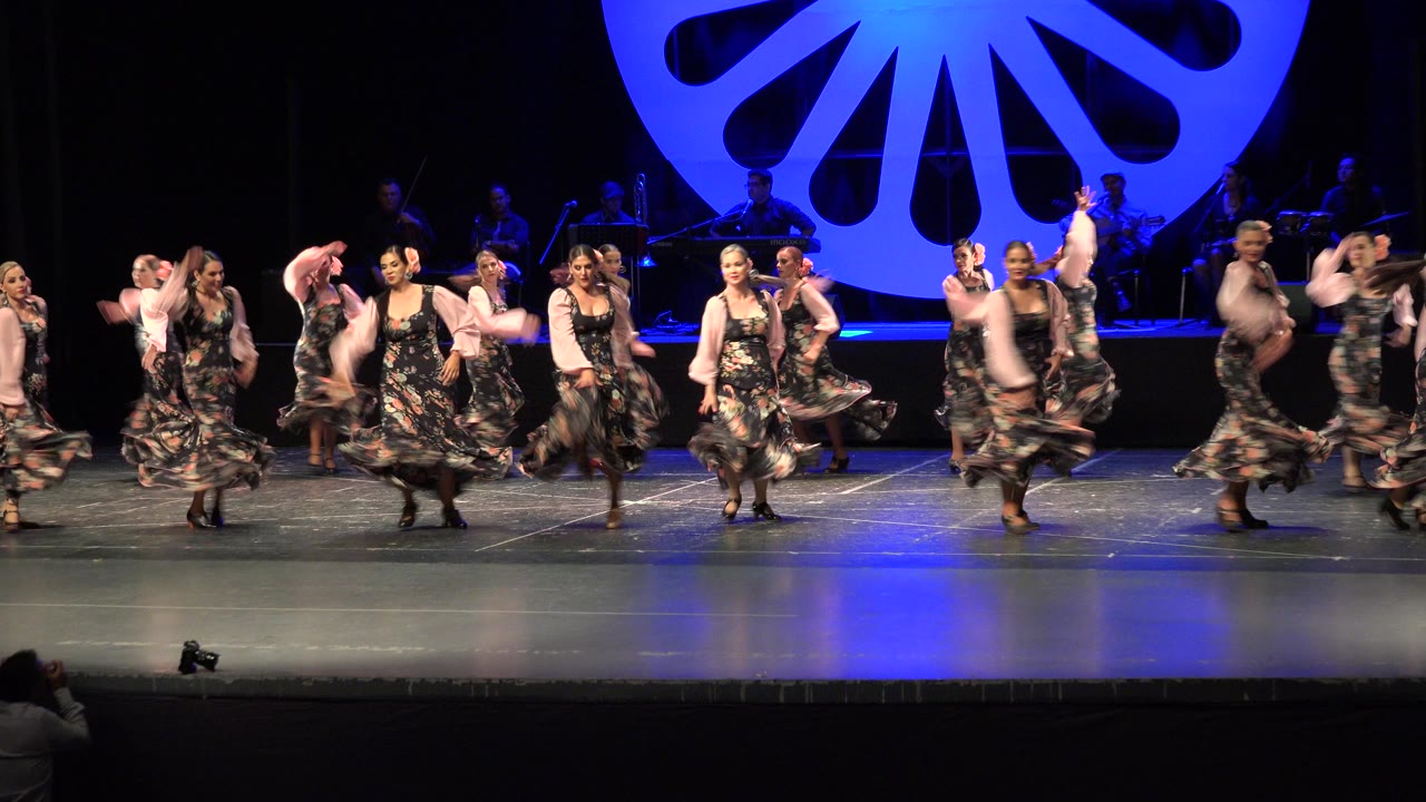 Wide stage view of women dancing in floral dresses under blue lighting.