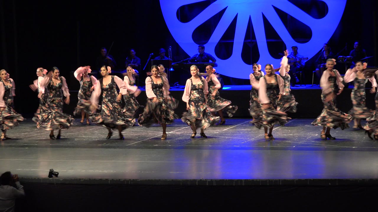Women performers in coordinated dresses continue the routine in Cabo San Lucas, Mexico, with several skirts flaring and the illuminated circular stage piece centered behind them.
