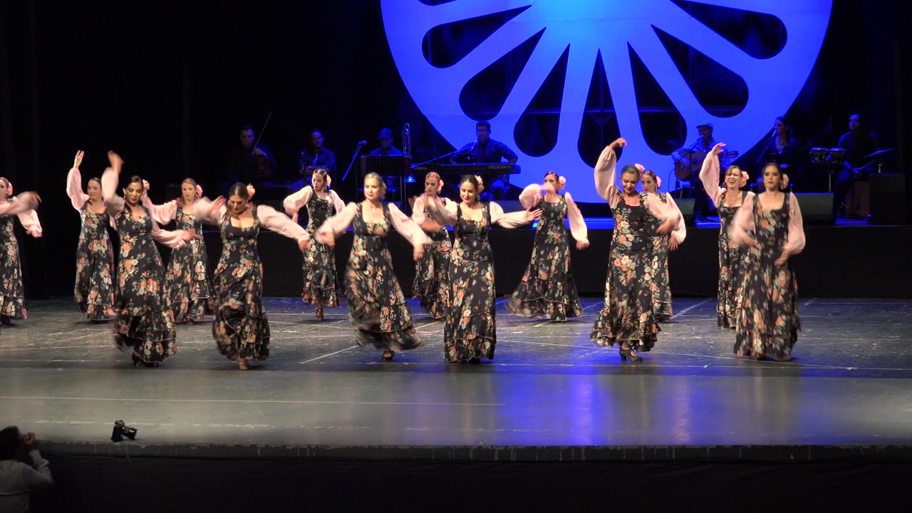 Women in matching floral dresses dance on a blue-lit stage.