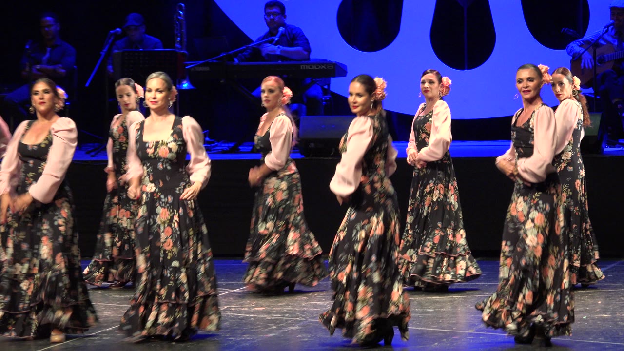 A line of adult women in matching floral costumes turns through the stage formation in Cabo San Lucas, Mexico, with blue backdrop lighting and dark instruments behind them.