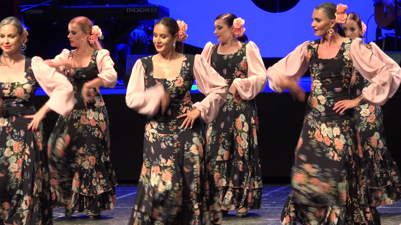 Women in black floral dresses with pale pink sleeves stand in formation on a blue-lit stage in Cabo San Lucas, Mexico, with performance equipment behind them.