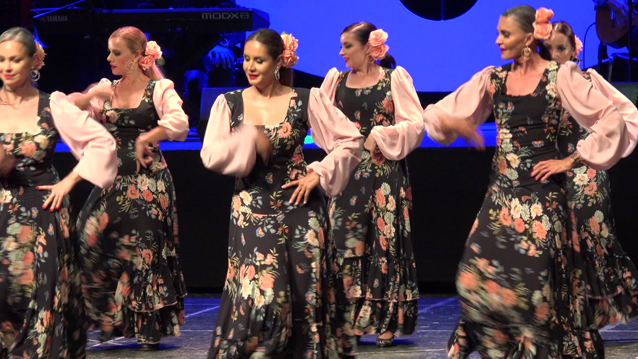 The group starts a coordinated movement in Cabo San Lucas, Mexico, with raised pink sleeves, floral skirts, and faces visible under blue stage light.