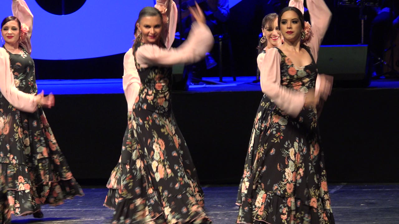 Adult women in black floral dresses with pale pink sleeves dance on a blue-lit stage in Cabo San Lucas, Mexico, with several faces visible near the front.