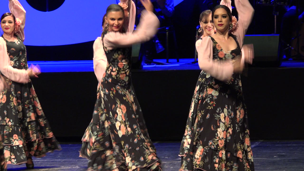 A front dancer in a black floral dress raises one arm while other women continue the synchronized stage dance behind her in Cabo San Lucas, Mexico.