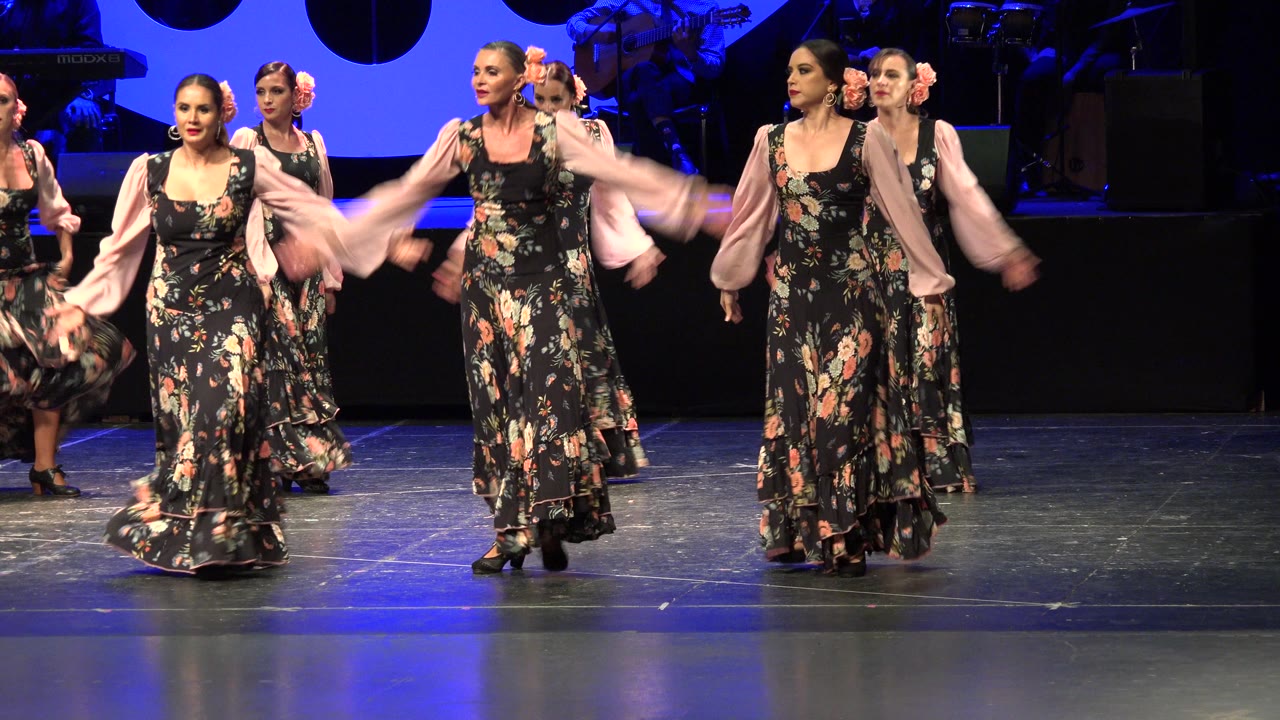 Women in floral dresses dance on a blue-lit stage.