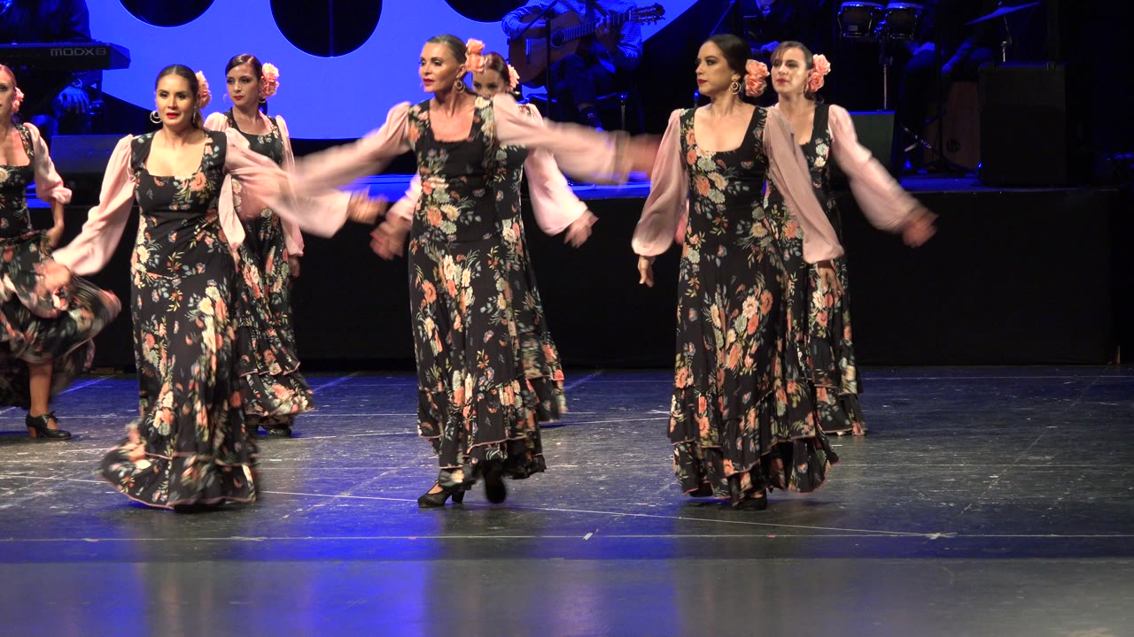 Several women in matching floral dresses continue the stage routine in Cabo San Lucas, with one dancer's pink sleeves blurred by motion near the center.