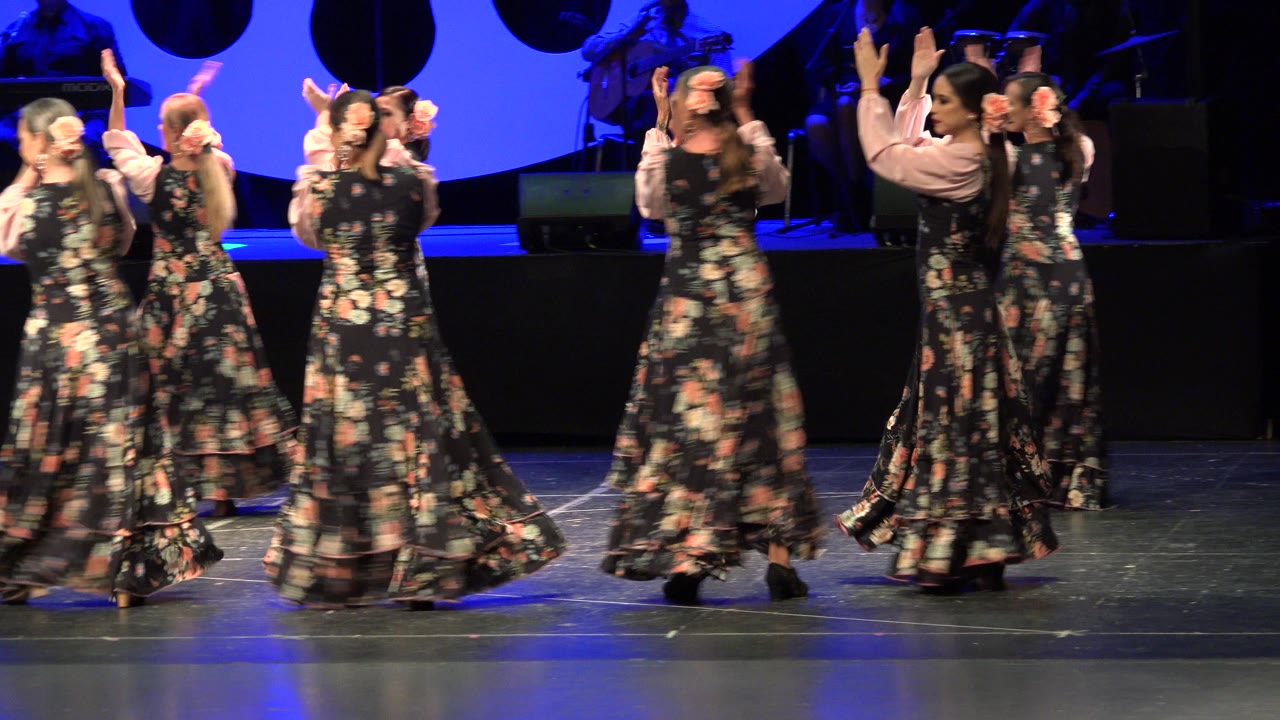 Women in floral dresses dance on a blue-lit theater stage.