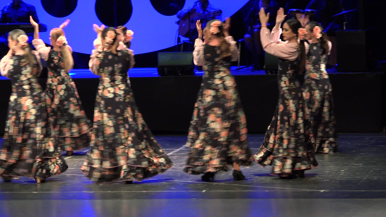 Several women in floral dresses turn and clap on the Cabo San Lucas, Mexico theater stage, with the blue backdrop and dark band area behind them.