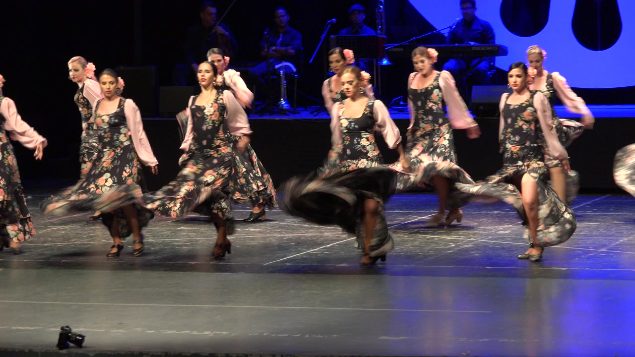 Women in floral dresses and pale sleeves perform on a stage in Cabo San Lucas, Mexico, with blue lighting and musicians in the dark background.